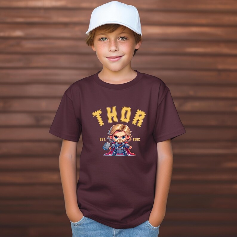 K&ouml;nnte beinhalten: Ein weinrotes T-Shirt mit dem Wort "THOR" in gelben Blockbuchstaben und einer Cartoon-Illustration von Thor. Die Illustration zeigt Thor mit einem Hammer und einem Umhang. Der Text "EST. 1962" steht darunter. Die Person tr&auml;gt eine wei&szlig;e Baseballkappe.