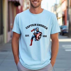 Pu&ograve; includere: T-shirt azzurra con grafica di Captain America. Il design include la scritta "CAPTAIN AMERICA" sopra un'immagine del personaggio, con "EST. 1918" sotto. La maglietta ha maniche corte.