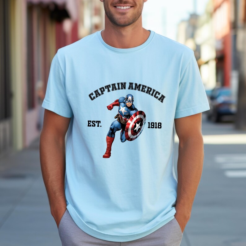 Pu&ograve; includere: T-shirt azzurra con grafica di Captain America. Il design include la scritta "CAPTAIN AMERICA" sopra un'immagine del personaggio, con "EST. 1918" sotto. La maglietta ha maniche corte.