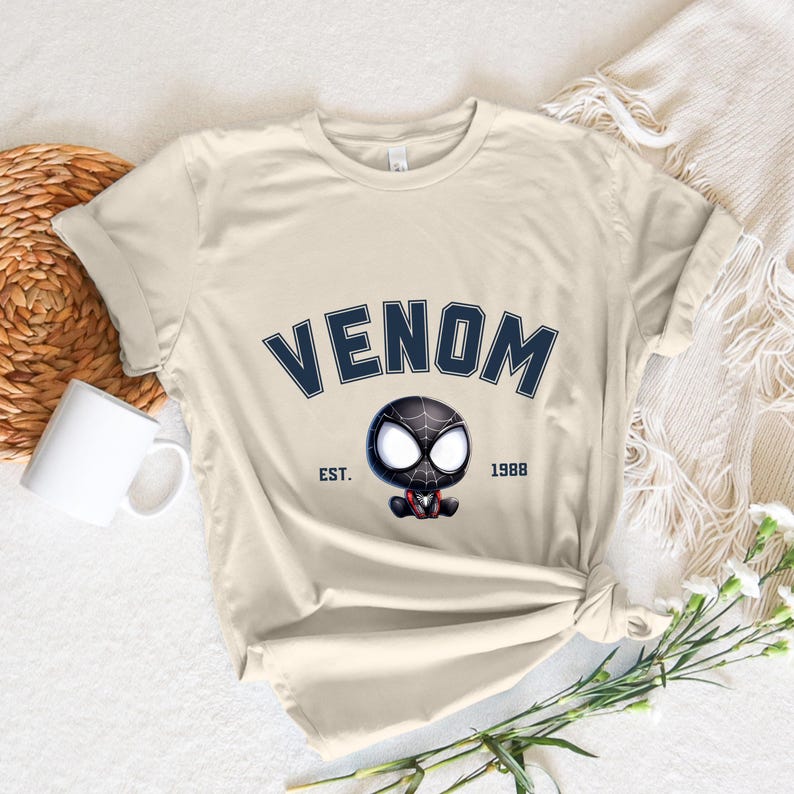 May include: Cream-colored t-shirt featuring the word "VENOM" in navy blue arched lettering above a cartoon Venom character. The text "EST. 1988" is below the character. The shirt is styled with a knot at the waist.
