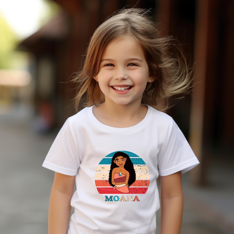 May include: A white t-shirt with a retro-style graphic of Moana from the Disney movie Moana. The graphic features Moana in a red and orange outfit with a blue ocean background.