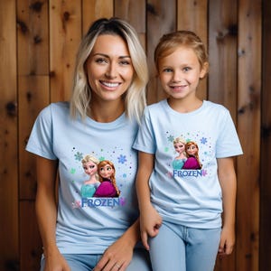 Puede incluir: Dos personas con camisetas de color azul claro con un estampado gr&aacute;fico de Frozen. El gr&aacute;fico presenta a Elsa y Anna de la pel&iacute;cula de Disney Frozen. El texto "FROZEN" est&aacute; impreso en las camisetas.