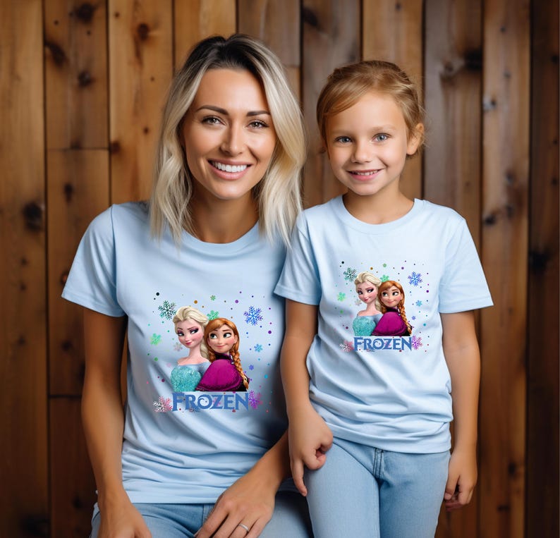 Puede incluir: Dos personas con camisetas de color azul claro con un estampado gr&aacute;fico de Frozen. El gr&aacute;fico presenta a Elsa y Anna de la pel&iacute;cula de Disney Frozen. El texto "FROZEN" est&aacute; impreso en las camisetas.