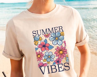 May include: A beige t-shirt with rolled-up sleeves featuring a floral design in a rectangular shape. The design includes pink, blue, and yellow flowers with the words "SUMMER VIBES" in navy blue.