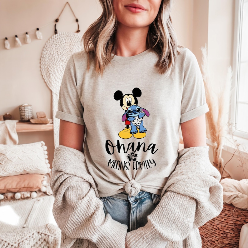 May include: A light gray t-shirt featuring Mickey Mouse and Stitch embracing. The text "Ohana Means Family" is printed below the characters. The shirt has short sleeves and a crew neck.