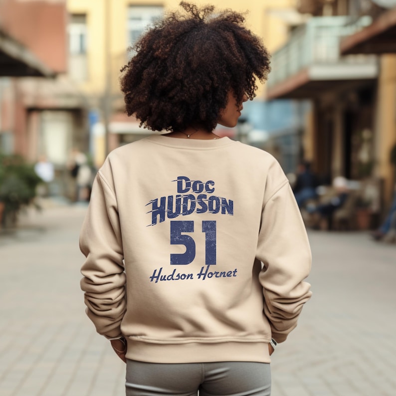 May include: A tan sweatshirt with the text "Doc Hudson 51 Hudson Hornet" in blue. The sweatshirt is a crew neck style and is made of a soft material. The image shows the back of the sweatshirt.