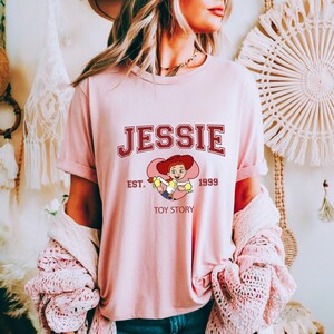 May include: A light pink t-shirt featuring the name "JESSIE" in maroon collegiate lettering. Below the name is a cartoon image of Jessie from Toy Story, with the text "EST. 1999 TOY STORY". The shirt has short sleeves and a crew neck.