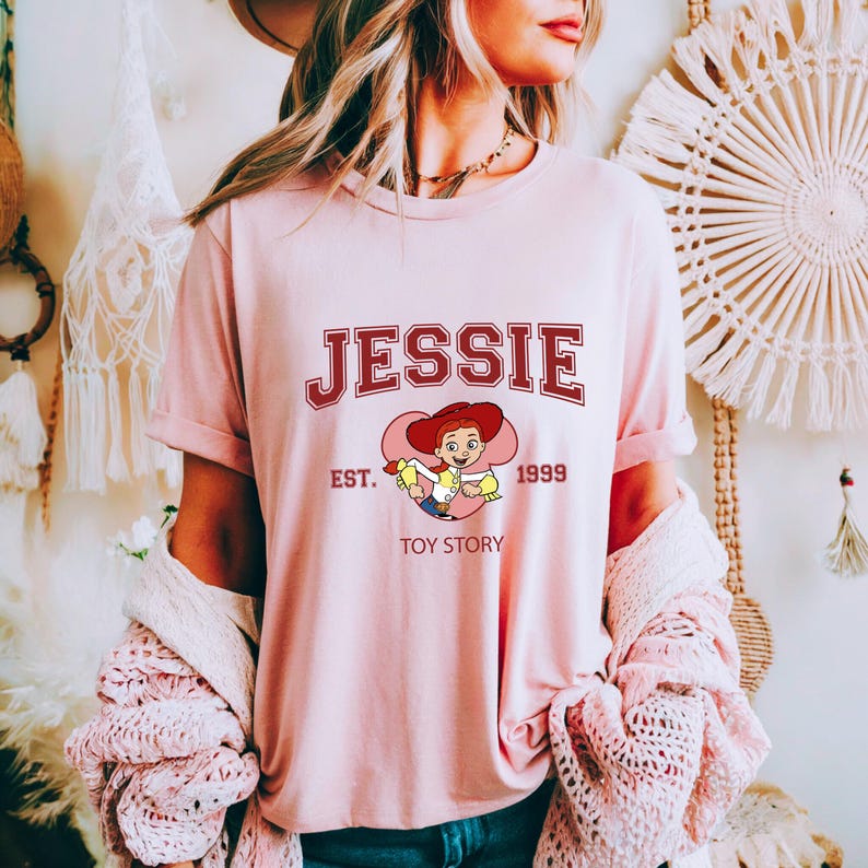 May include: A light pink t-shirt featuring the name "JESSIE" in maroon collegiate lettering. Below the name is a cartoon image of Jessie from Toy Story, with the text "EST. 1999 TOY STORY". The shirt has short sleeves and a crew neck.
