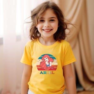 Puede incluir: Una camiseta amarilla con un gr&aacute;fico de estilo vintage de Ariel de La Sirenita. El gr&aacute;fico presenta a Ariel con cabello rojo y un sujetador de concha azul, sobre un fondo de puesta de sol colorido. El texto "ARIEL" est&aacute; impreso debajo del gr&aacute;fico.