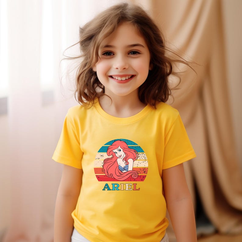 Puede incluir: Una camiseta amarilla con un gr&aacute;fico de estilo vintage de Ariel de La Sirenita. El gr&aacute;fico presenta a Ariel con cabello rojo y un sujetador de concha azul, sobre un fondo de puesta de sol colorido. El texto "ARIEL" est&aacute; impreso debajo del gr&aacute;fico.