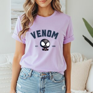 May include: A light purple t-shirt featuring the word "VENOM" in navy blue arched text above a cartoon Venom character. The text "EST. 1988" is below the character. The shirt has rolled-up sleeves.