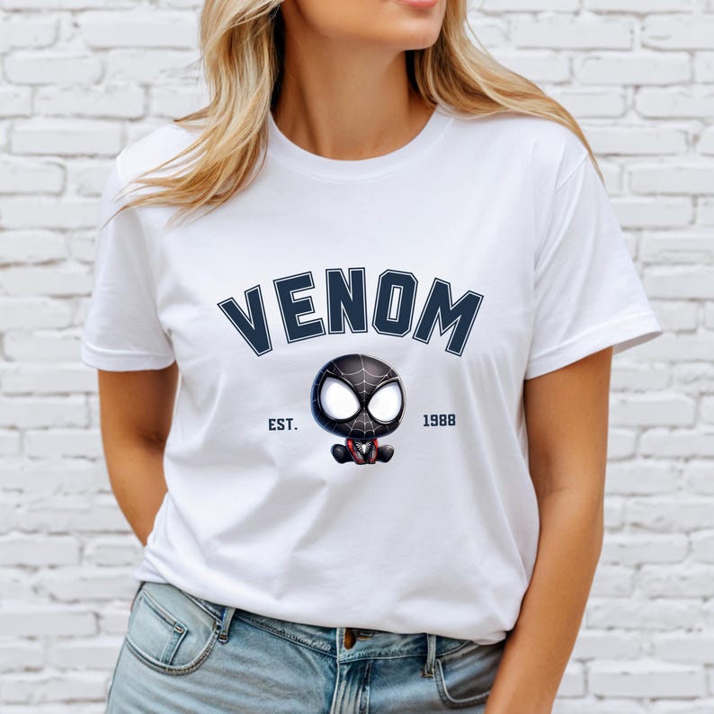 May include: White t-shirt featuring the word "VENOM" in navy blue arched lettering above a cartoon Venom character. The character is black with white eyes and a red mouth. The text "EST. 1988" is below the character.