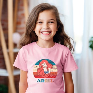 Puede incluir: Camiseta rosa con un gr&aacute;fico de estilo vintage de Ariel de La Sirenita. El gr&aacute;fico presenta una puesta de sol roja, naranja, amarilla y azul con el retrato de Ariel en el centro. El texto "ARIEL" est&aacute; impreso debajo del gr&aacute;fico.