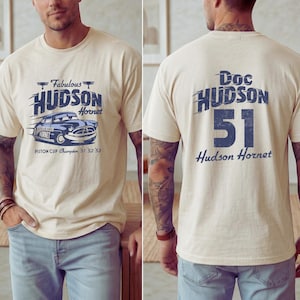 May include: Cream-colored t-shirt featuring a blue graphic of a vintage car with the text "Fabulous Hudson Hornet" and "Piston Cup Champion '51 '52 '53" on the front. The back of the shirt displays "Doc Hudson 51 Hudson Hornet" in blue.