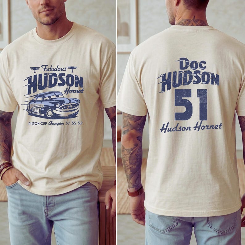 May include: Cream-colored t-shirt featuring a blue graphic of a vintage car with the text "Fabulous Hudson Hornet" and "Piston Cup Champion '51 '52 '53" on the front. The back of the shirt displays "Doc Hudson 51 Hudson Hornet" in blue.
