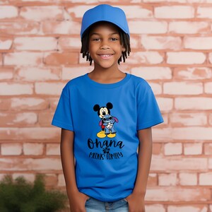 May include: A blue t-shirt featuring Mickey Mouse and Stitch hugging, with the text "Ohana Means Family" in black. The child is wearing a blue baseball cap. The background is a brick wall.