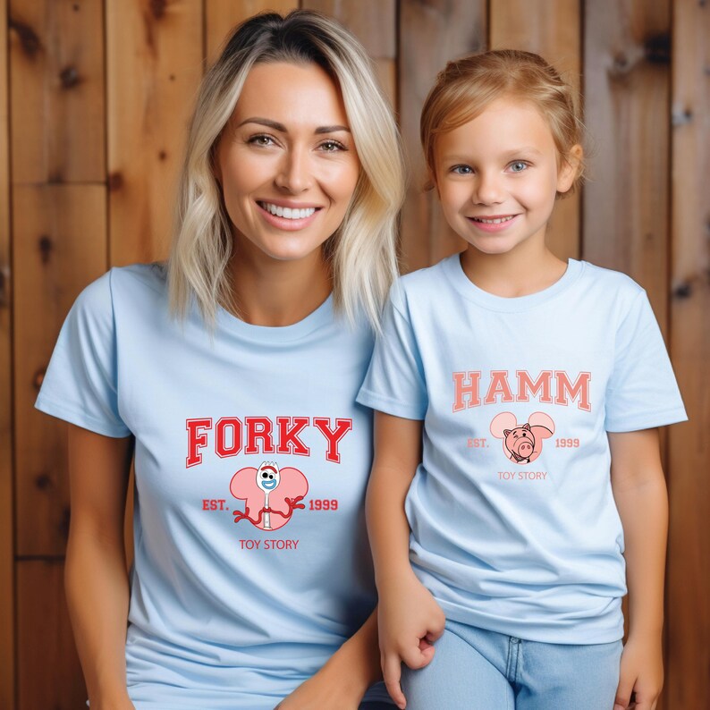 May include: Two light blue t-shirts with matching graphics. One shirt features the word "FORKY" in red, with a pink character illustration and the text "EST. 1999 TOY STORY." The other shirt says "HAMM" in pink, with a pig illustration.