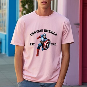 Pu&ograve; includere: Una maglietta rosa chiaro con una grafica di Captain America. Il testo "CAPTAIN AMERICA" &egrave; sopra l'immagine, e "EST. 1918" sotto. L'illustrazione mostra il supereroe nel suo costume iconico, con il suo scudo.