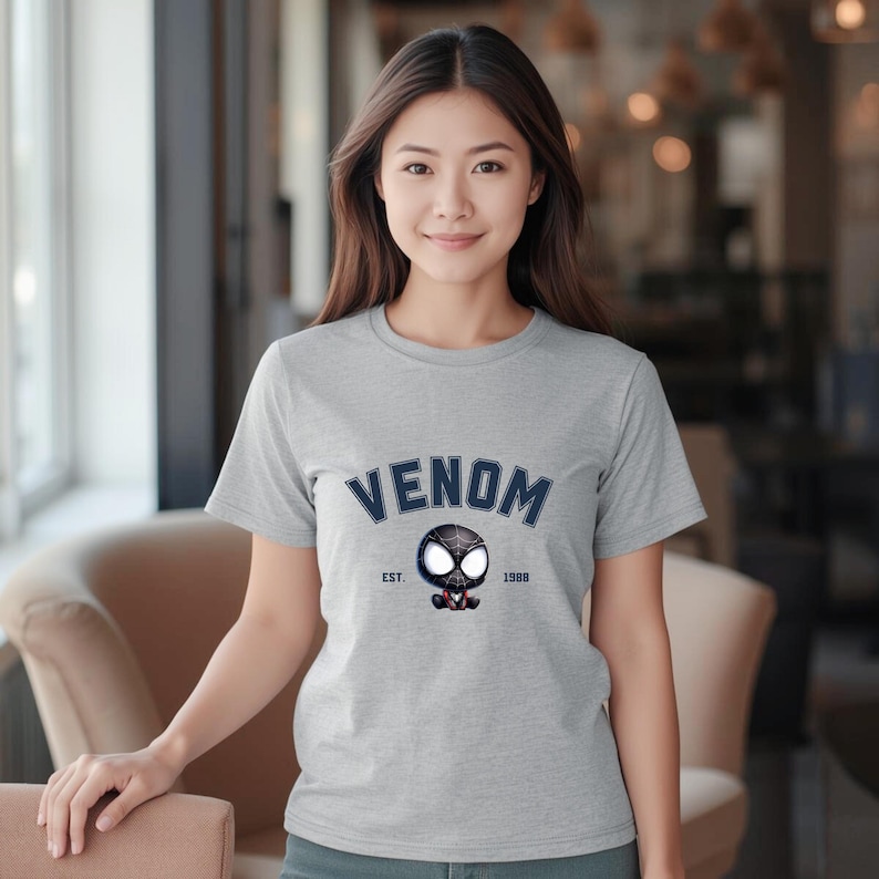 May include: A light gray t-shirt featuring the word "VENOM" in navy blue arched text above a cartoon graphic of Venom. The graphic includes the text "EST. 1988". The shirt is short-sleeved.