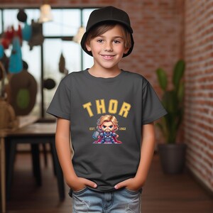 K&ouml;nnte beinhalten: Ein anthrazitfarbenes T-Shirt mit dem Wort "THOR" in gelben Blockbuchstaben und einer Cartoon-Illustration von Thor. Das T-Shirt hat auch den Text "EST. 1962". Das T-Shirt wird von einem Kind getragen.