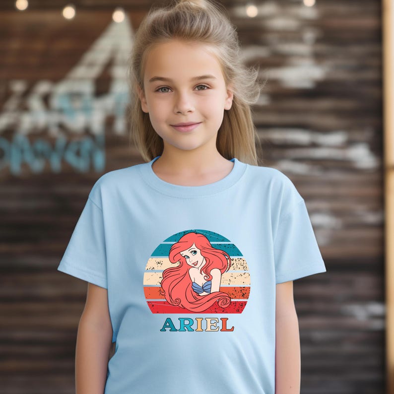 Puede incluir: Camiseta azul claro con un gr&aacute;fico de puesta de sol retro con Ariel de La Sirenita. El gr&aacute;fico incluye una puesta de sol roja, naranja, amarilla y azul con Ariel en el centro. El texto "ARIEL" est&aacute; impreso debajo del gr&aacute;fico.
