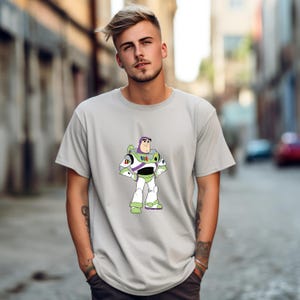 May include: A light gray t-shirt featuring a cartoon image of Buzz Lightyear from Toy Story. The character is depicted in his signature white, green, and purple spacesuit. The shirt has short sleeves and a crew neck.
