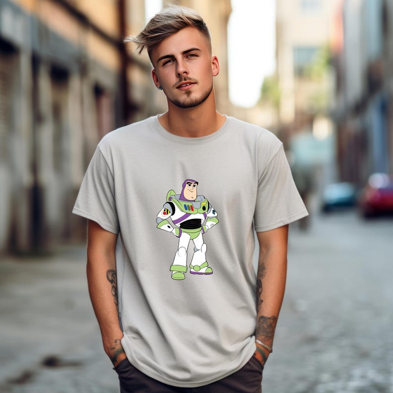 May include: A light gray t-shirt featuring a cartoon image of Buzz Lightyear from Toy Story. The character is depicted in his signature white, green, and purple spacesuit. The shirt has short sleeves and a crew neck.