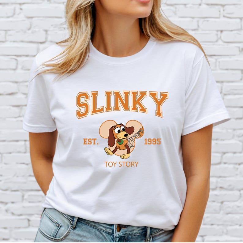 May include: White t-shirt featuring the word "SLINKY" in orange block letters above a cartoon image of Slinky Dog from Toy Story. The shirt also includes "EST. 1995" and "TOY STORY" in orange text.