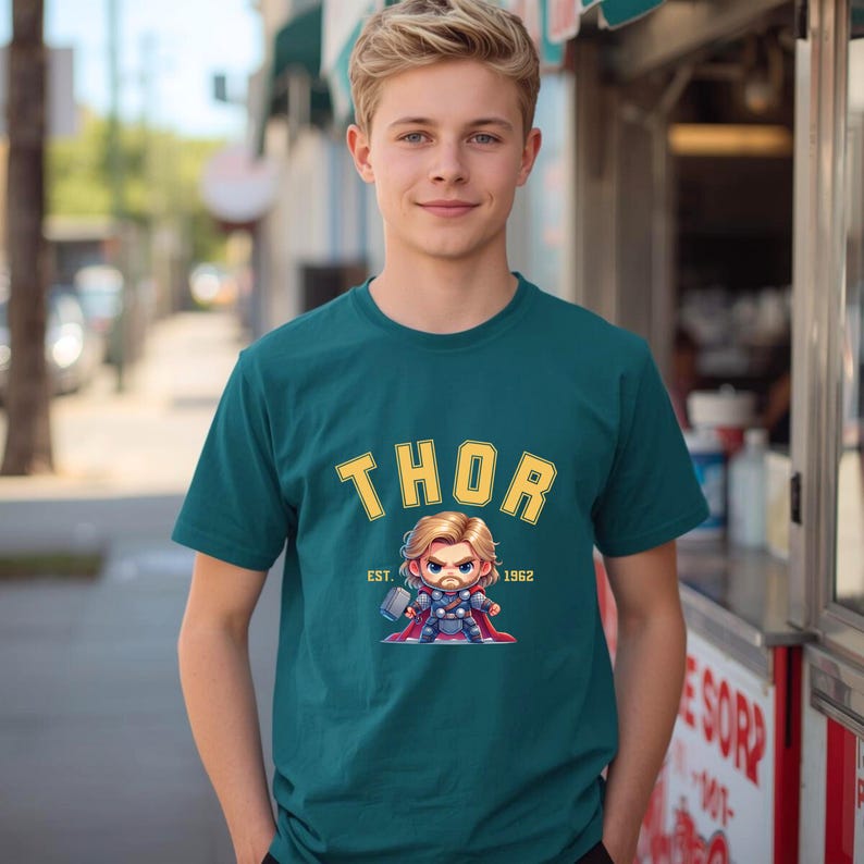 K&ouml;nnte beinhalten: T&uuml;rkisfarbenes T-Shirt mit gelbem, bogenf&ouml;rmigem "THOR"-Schriftzug und einer Cartoon-Illustration von Thor mit einem Hammer. Der Text "EST. 1962" steht darunter. Das T-Shirt wird von einer Person getragen.