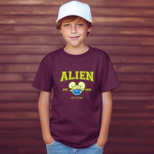 May include: A maroon t-shirt with the word "ALIEN" in yellow block letters and a cartoon alien graphic. The shirt also features the text "EST. 2010" and "TOY STORY". The person is wearing a white baseball cap.