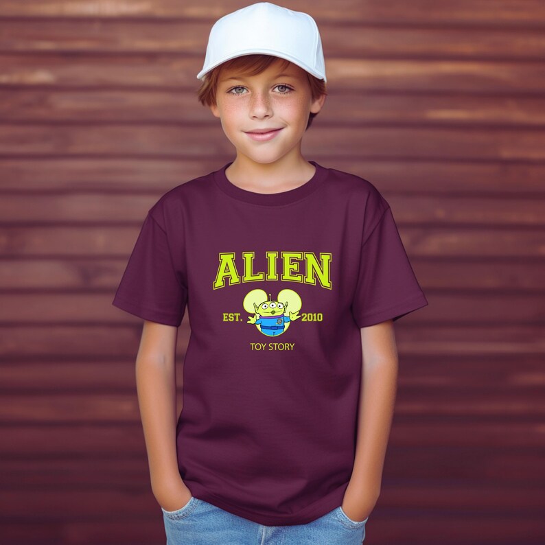 May include: A maroon t-shirt with the word "ALIEN" in yellow block letters and a cartoon alien graphic. The shirt also features the text "EST. 2010" and "TOY STORY". The person is wearing a white baseball cap.