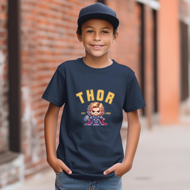 May include: Navy blue t-shirt featuring a cartoon illustration of Thor holding a hammer. The word "THOR" is printed in yellow arched lettering above the image, with "EST. 1962" below. The shirt is worn by a person wearing a black baseball cap.