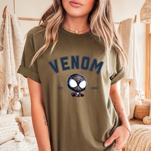 May include: Olive green t-shirt with the word "VENOM" in navy blue block letters above a cartoon Venom character. The text "EST. 1988" is below the character. The shirt has rolled-up sleeves.