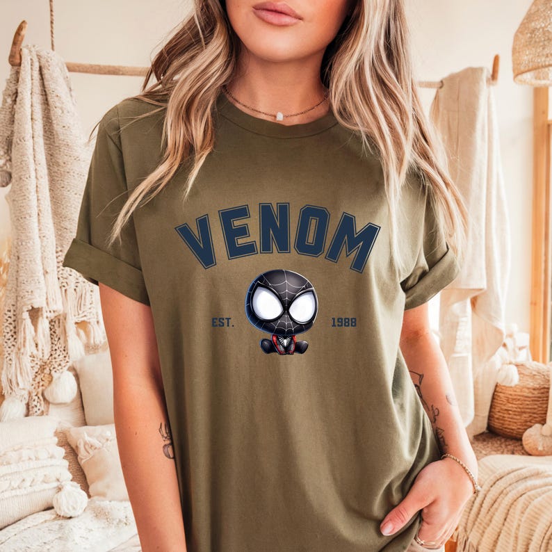 May include: Olive green t-shirt with the word "VENOM" in navy blue block letters above a cartoon Venom character. The text "EST. 1988" is below the character. The shirt has rolled-up sleeves.