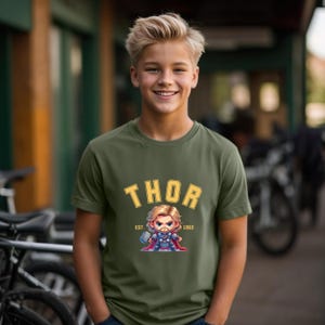 K&ouml;nnte beinhalten: Olivgr&uuml;nes T-Shirt mit dem Wort "THOR" in goldfarbenen Buchstaben und einer Cartoon-Illustration von Thor. Darunter steht "EST. 1962". Das T-Shirt hat einen Rundhalsausschnitt und ist aus weichem Material.