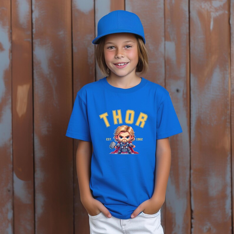 K&ouml;nnte beinhalten: Ein blaues T-Shirt mit dem Wort "THOR" in gelben Buchstaben und einem Cartoon-Bild von Thor. Das Shirt hat auch den Text "EST. 1962". Die Person auf dem Bild tr&auml;gt eine blaue Baseballkappe und eine wei&szlig;e Hose.