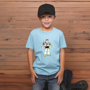May include: A light blue t-shirt featuring a cartoon image of Buzz Lightyear from Toy Story. The character is depicted in his signature space ranger suit, with green, white, and purple accents. The shirt is worn by a child wearing a black baseball cap and blue jeans.
