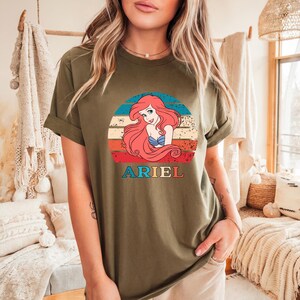 Puede incluir: Una camiseta verde con un gr&aacute;fico de estilo vintage de Ariel, la Sirenita, con el texto "ARIEL" debajo del gr&aacute;fico.