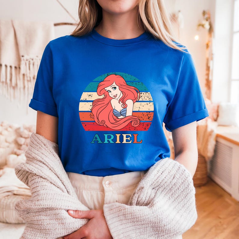Puede incluir: Una camiseta azul real con un gr&aacute;fico de estilo vintage de Ariel, la Sirenita, con el texto "ARIEL" debajo del gr&aacute;fico.