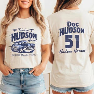 May include: Beige t-shirt featuring a blue graphic of a Hudson Hornet car with the text "Fabulous Hudson Hornet" and "Piston Cup Champion '51 '52 '53". The back of the shirt displays "Doc Hudson 51 Hudson Hornet" in blue.