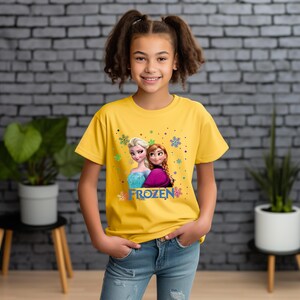 Puede incluir: Una camiseta amarilla con una imagen de Elsa y Anna de la pel&iacute;cula Frozen. La camiseta tiene la palabra "FROZEN" impresa.