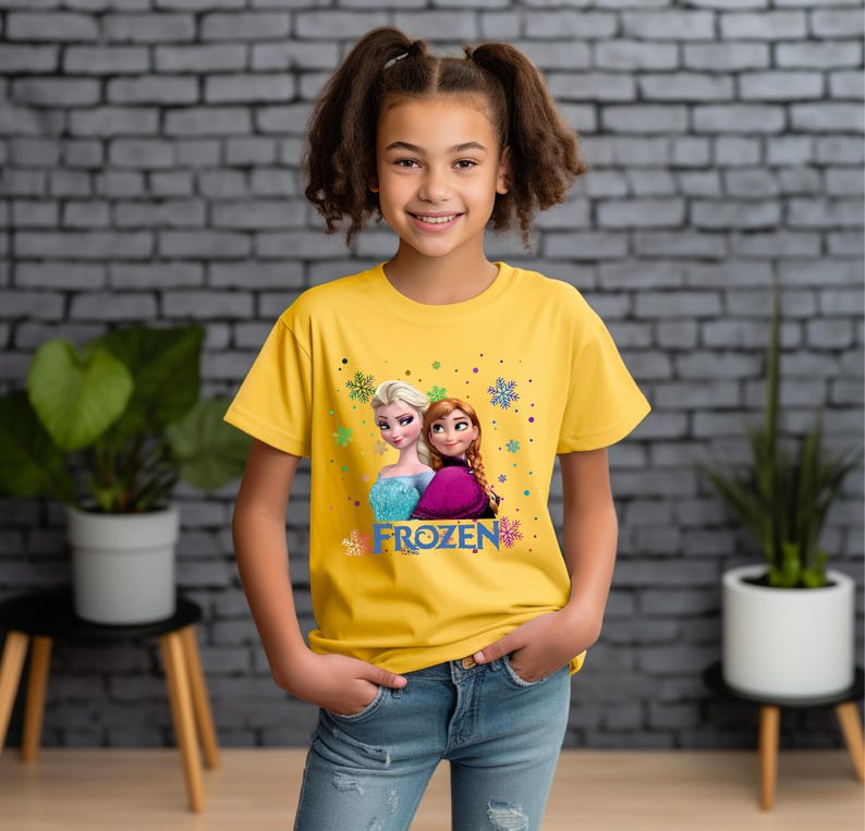 Puede incluir: Una camiseta amarilla con una imagen de Elsa y Anna de la pel&iacute;cula Frozen. La camiseta tiene la palabra "FROZEN" impresa.