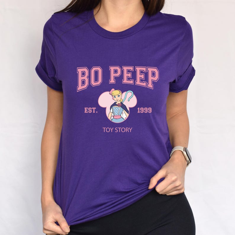 May include: A purple t-shirt featuring the text "BO PEEP" in pink, with "EST. 1999" and "TOY STORY" below. The design includes an illustration of Bo Peep from Toy Story. The shirt has rolled-up sleeves.