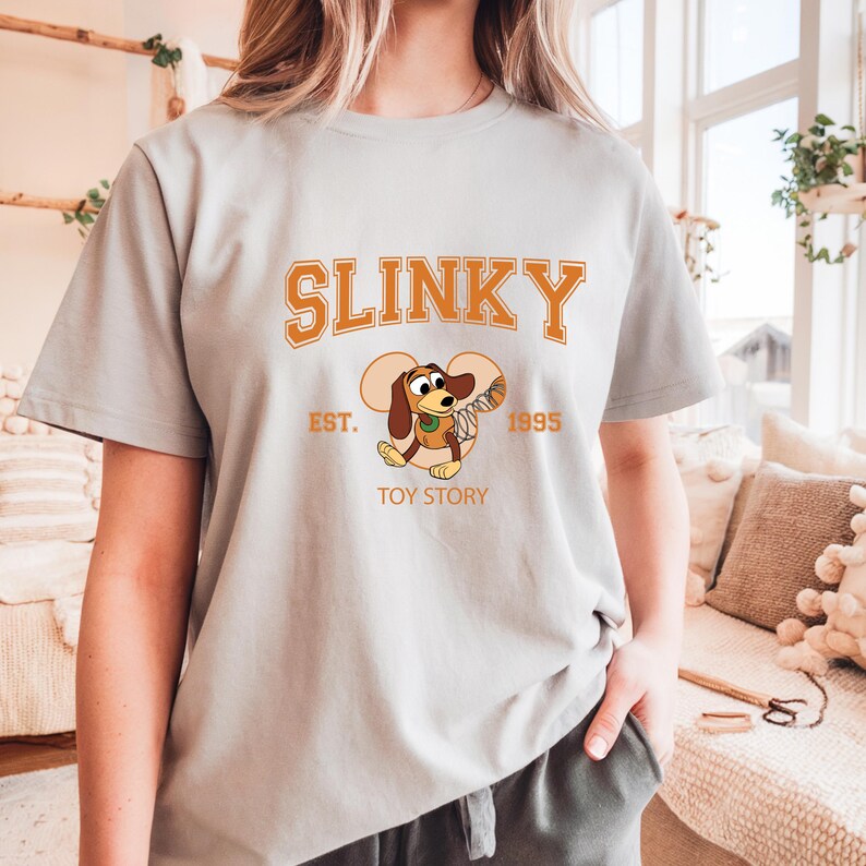 May include: A light gray t-shirt featuring the word "SLINKY" in orange block letters above a cartoon image of Slinky Dog from Toy Story. The shirt also includes the text "EST. 1995" and "TOY STORY" in orange.