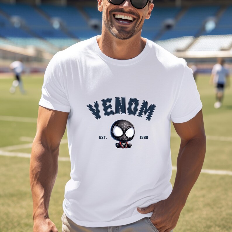 May include: White t-shirt featuring the word "VENOM" in navy blue arched text above a cartoon graphic of Venom's head. The graphic includes the text "EST. 1988". The shirt is a classic crew neck style.