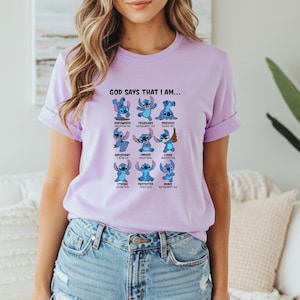 May include: A light purple t-shirt featuring a graphic of the character Stitch from Lilo & Stitch. The graphic includes the text "GOD SAYS THAT I AM..." and various positive affirmations like "EMPOWERED" and "BRAVE".