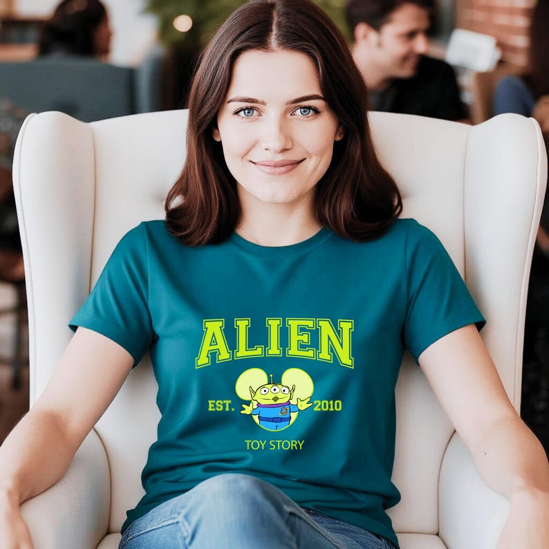 May include: Teal t-shirt with the word "ALIEN" in bright green block letters. Below the word "ALIEN" is a cartoon alien character with the text "EST. 2010 TOY STORY". The shirt is being worn by a person.