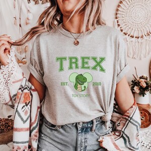 May include: A gray t-shirt with a green "TREX" graphic, a cartoon dinosaur, and the text "EST. 2010 TOY STORY." The shirt is a crew neck and has rolled-up sleeves. The shirt is worn with blue jeans.