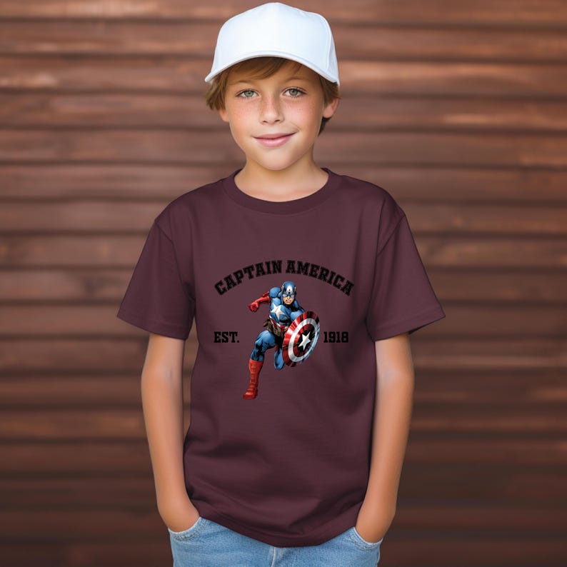 Pu&ograve; includere: Una maglietta bordeaux con una grafica di Captain America e la scritta "CAPTAIN AMERICA EST. 1918". La grafica raffigura Captain America con il suo scudo. La persona nell'immagine indossa un berretto da baseball bianco.