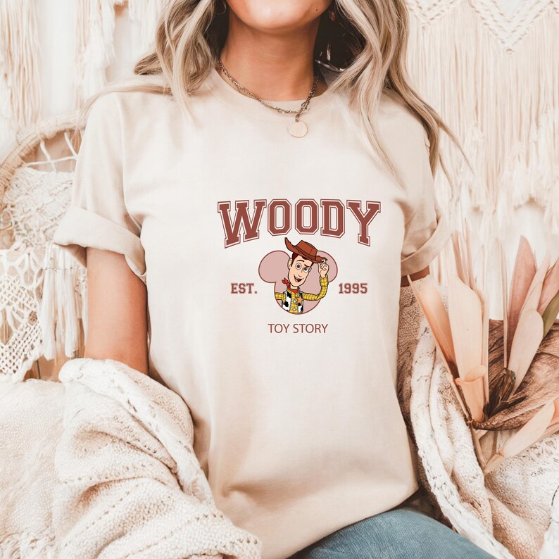 May include: Beige t-shirt featuring the word "WOODY" in maroon, with "EST. 1995 TOY STORY" below a cartoon image of Woody from Toy Story. The shirt has short sleeves and a crew neck.